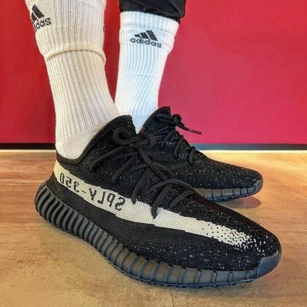 Black knit sneakers with a white SPLY-350 stripe, worn with white Adidas socks on a wooden floor.