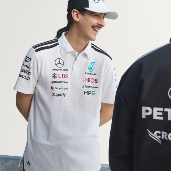 Smiling man in a white polo with sponsor logos (Mercedes, Petronas, UBS) and a two-tone cap, standing next to a dark-clad person.