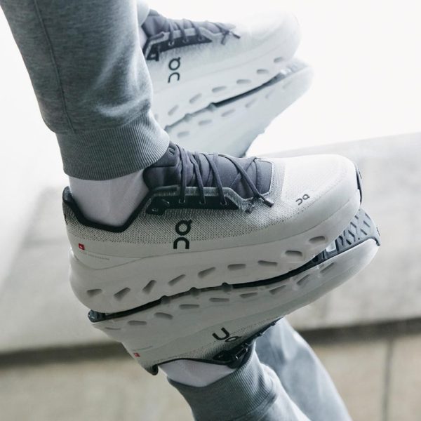 Close-up of white and gray On running shoes being worn, mid-step with gray joggers visible behind them.