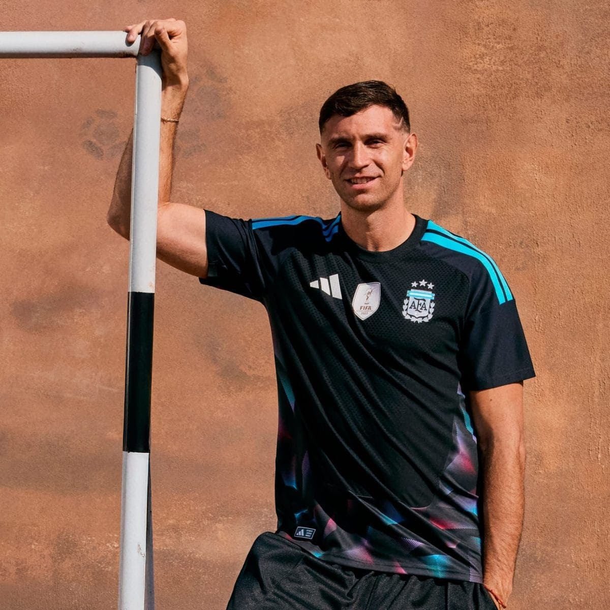 Man in a dark training jersey with blue stripes leans on a white-and-black pole, stands against a brown textured wall and smiles at the camera.