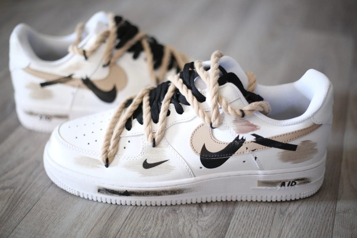 White sneakers with beige and black swoosh logos and tangled beige laces on a wooden floor.