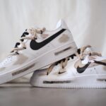 White Nike sneakers with beige paint-splatter accents and a black swoosh, stacked on a light floor.
