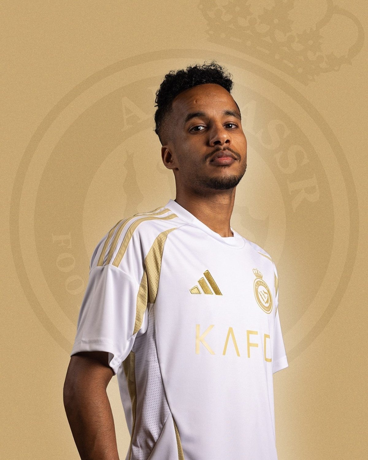 Portrait of a man in a white and gold football jersey with a crest, posing against a beige background with a circular emblem behind him.