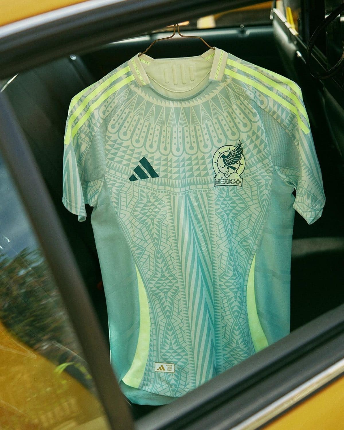 Mexico national team jersey with teal geometric pattern, hanging on a hanger inside a car, Adidas logo visible on chest goalkeepers style.