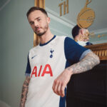Man in Tottenham jersey leaning on a piano, visible in a gold-framed mirror reflection.