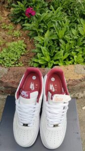 Pair of white Nike Air Force 1 sneakers with red inner lining resting on a dark mat outdoors beside green plants and a pink flower in a garden edge.