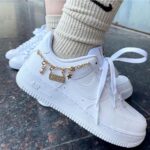 White Nike Air Force 1 sneakers with a gold chain anklet and charms on a beige socked ankle, outdoor on a pavement.