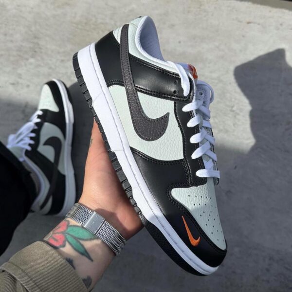 Close-up of a Nike Dunk Low sneaker in white, black, and mint with an orange Swoosh, held in a hand showing the midfoot and sole.