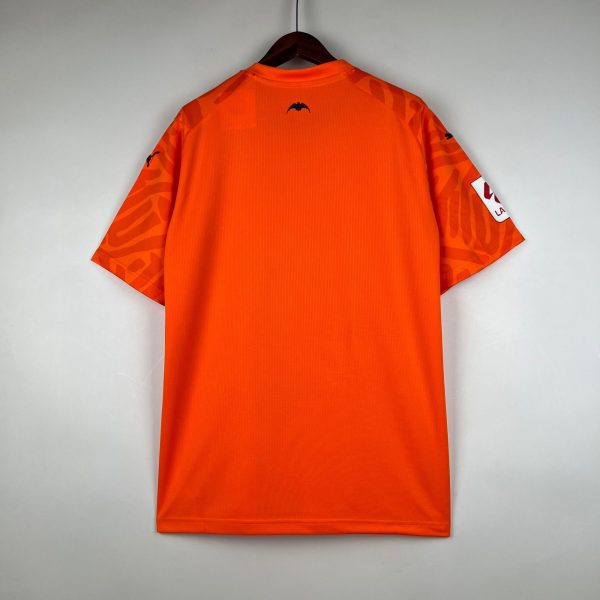 Orange sports jersey hanging on a wooden hanger against a gray wall, viewed from the back; small dark bat logo at the collar.
