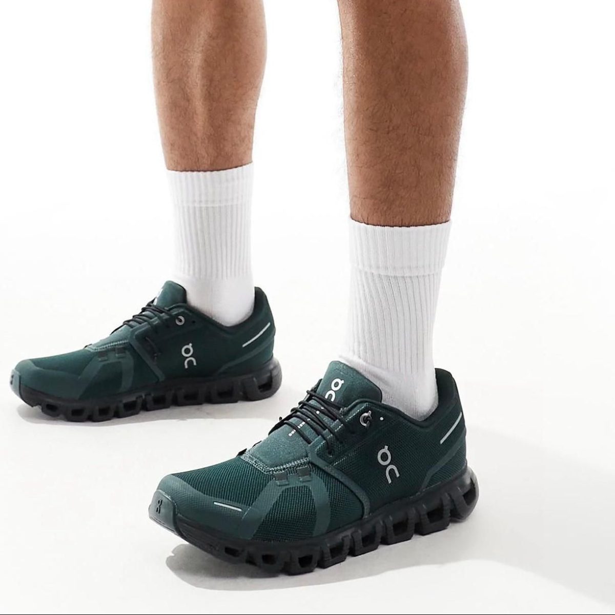 Pair of dark green running shoes with thick, segmented black soles and white crew socks standing on a white background.