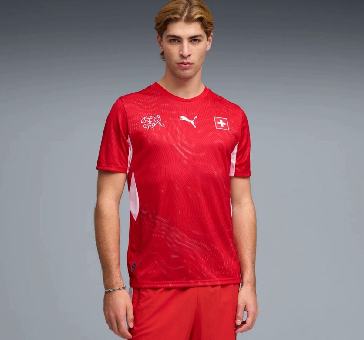 Young man wearing a red Swiss national football jersey with white logos, standing against a gray gradient background, facing the camera.