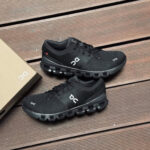 Pair of black On running shoes displayed on a wooden deck beside a shoe box.