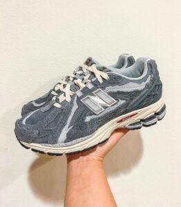 Gray New Balance 990 sneaker held in a hand against a pale wall, displaying its side and logo.