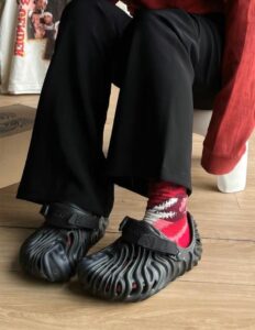 9bb00bf2-7bc5-4150-9f89-ade10bc7b336 | UAE SPORT STORE Close-up of a person wearing black wavy-patterned clogs with straps and red socks, sitting on a wooden floor.