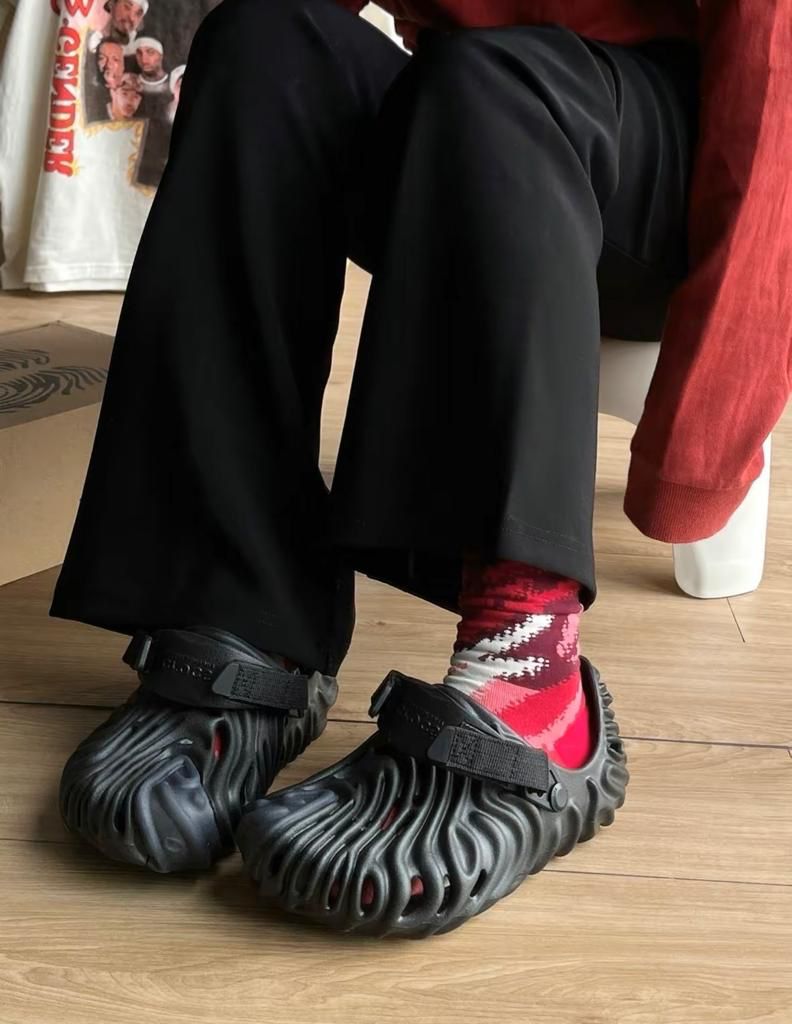 9bb00bf2-7bc5-4150-9f89-ade10bc7b336 | UAE SPORT STORE Close-up of a person wearing black wavy-patterned clogs with straps and red socks, sitting on a wooden floor.