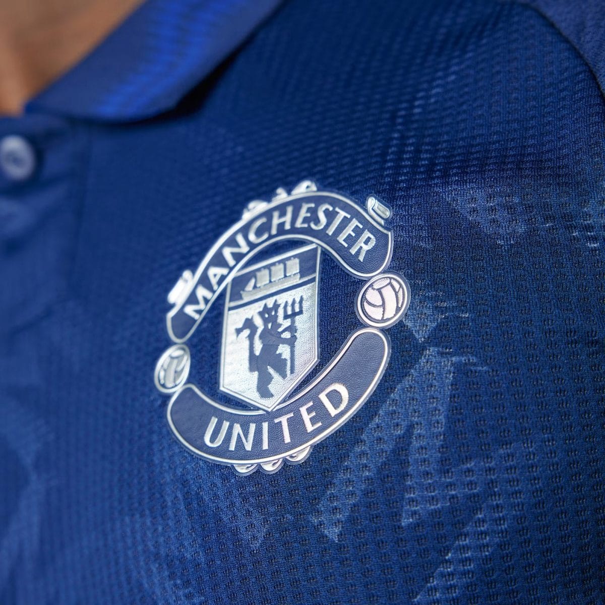 Close-up of the Manchester United badge on a blue jersey fabric, showing the emblem and lettering.