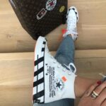 White high-top Converse sneaker with black diagonal stripes and orange sole, worn with jeans beside a patterned suitcase on a wooden floor.