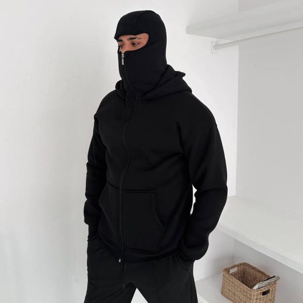 Person wearing a black balaclava and zip-up hoodie with hands in pockets, standing in a white room.