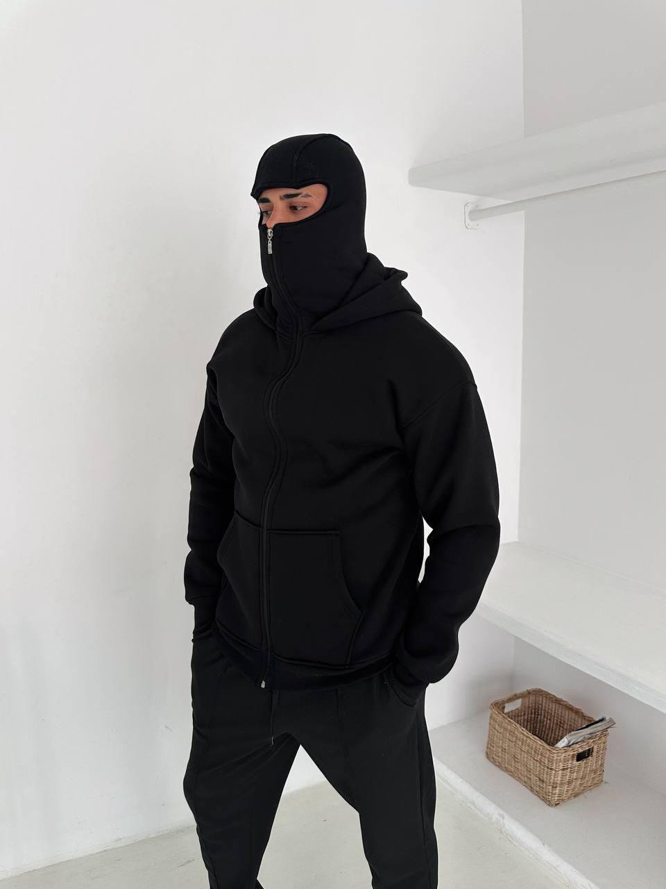 9c6b1411-3197-4c48-8dab-ddaf4edc2aa9 | UAE SPORT STORE Person wearing a black balaclava and zip-up hoodie with hands in pockets, standing in a white room.