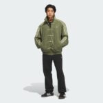 Young man posing in an olive-green duffle jacket with white stripes, paired with black pants and Adidas sneakers.