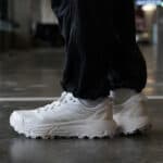 White athletic sneakers with chunky soles on a person wearing black pants, standing indoors on a glossy floor.