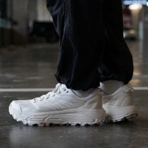 White athletic sneakers with chunky soles on a person wearing black pants, standing indoors on a glossy floor.