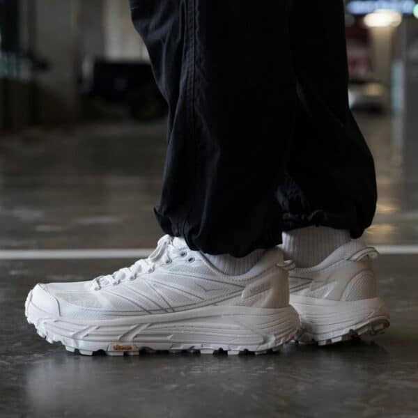 White athletic sneakers with chunky soles on a person wearing black pants, standing indoors on a glossy floor.