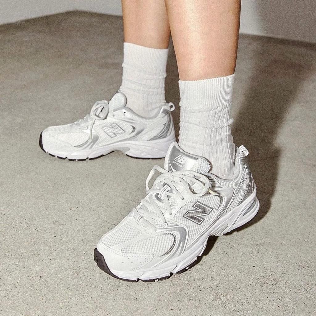 9cb6cce3-e409-4290-a6cc-f7cea7c9338d Close-up of white New Balance running shoes on a person’s feet, wearing white crew socks, standing on a concrete floor.