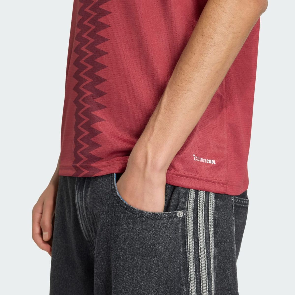 Side view of a person wearing a red athletic shirt with a zigzag stripe and a small ClimaCool logo, paired with dark jeans with white vertical stripes near the pocket.
