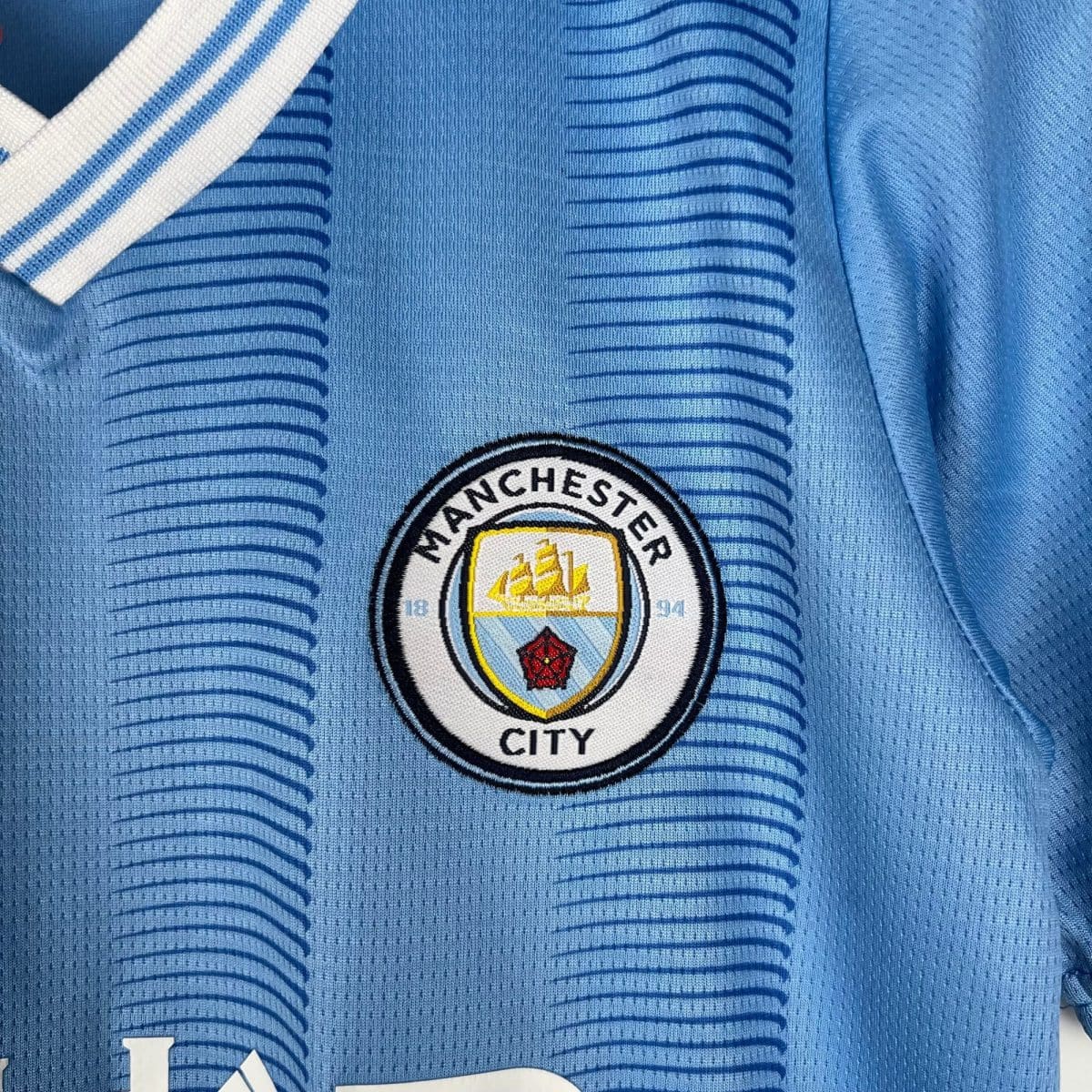 Close-up of a light blue Manchester City football jersey fabric with a round crest patch showing club emblem and skyline details.