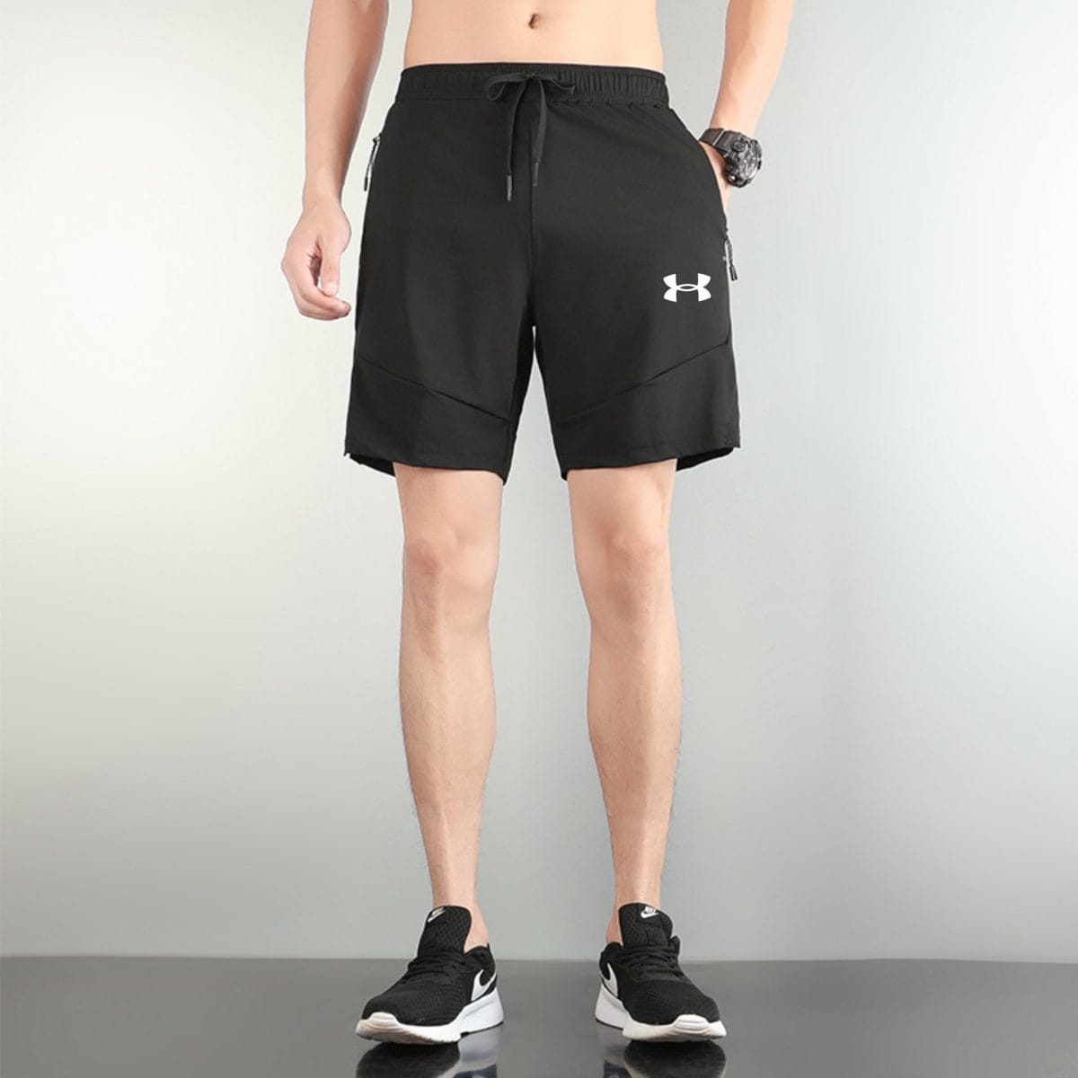 Person wearing black athletic shorts with a drawstring, standing on a reflective floor in black and white athletic shoes (Under Armour logo visible).