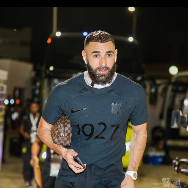 Athletic man with a beard walking at night, wearing a dark shirt with '1927' and a crest, headphones in, holding a phone and a brown checkered bag under his arm.
