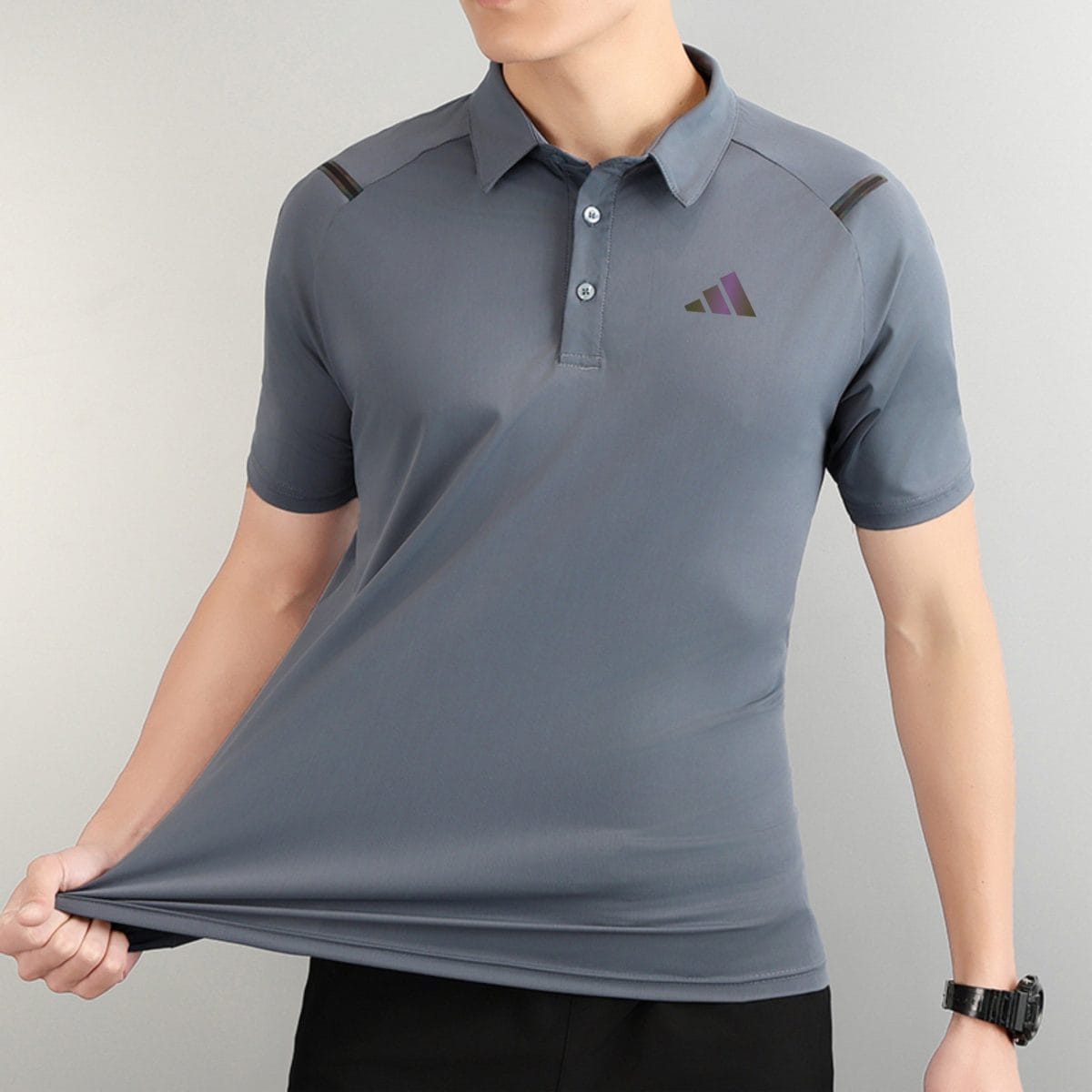 Person wearing a gray polo shirt with a small three-triangle logo on the left chest, pulling the hem downward.