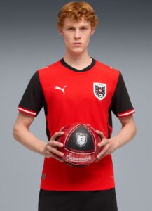 Young male soccer player in a red and black jersey holds a red soccer ball toward the camera, facing forward.