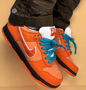 Orange Nike SB sneakers with turquoise laces and white swoosh, paired with brown pants, hanging side-by-side against a tan background.