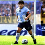 Soccer player in light blue and white jersey with number 10 dribbling a ball on a grass field during a match, wearing a captain's armband.