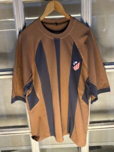 Brown sports jersey with dark navy vertical panels, hanging on a wooden hanger; small shield-style club crest on the left chest featuring red-and-white stripes and a blue field.