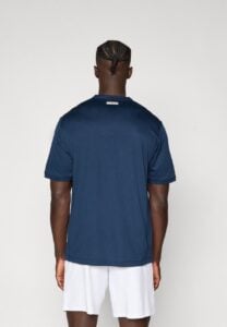 Back view of a man wearing a navy T-shirt and white shorts, with braided cornrow hairstyle, standing against a light wall.