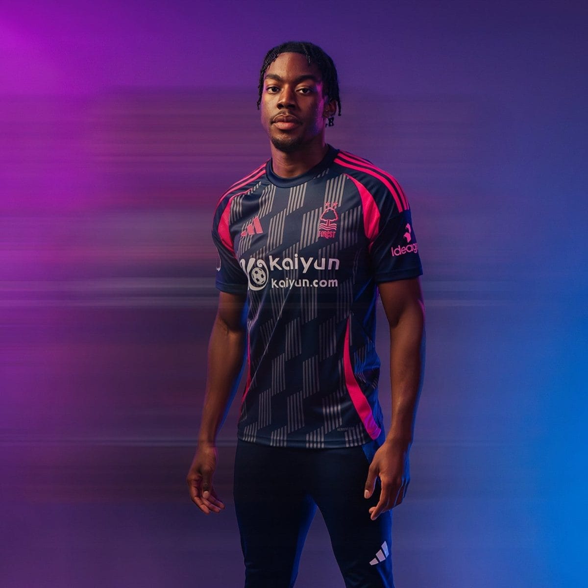 Portrait of a man in a dark navy sports jersey with pink accents, standing against a purple-blue gradient background with motion blur.