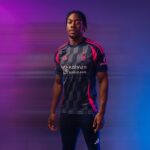 Portrait of a man in a dark navy sports jersey with pink accents, standing against a purple-blue gradient background with motion blur.