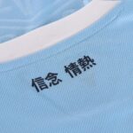 Close-up of light blue fabric with a white collar and black Japanese characters reading 信念 and 情熱 along the seam, conveying belief and passion.