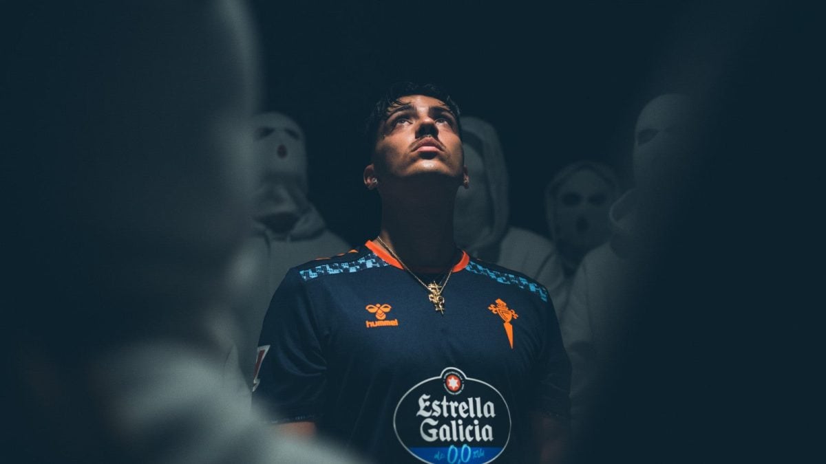 Young man in a dark blue sports jersey with orange logos, glowing under dramatic light, surrounded by blurred masked figures in the background.