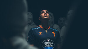 Young man in a dark blue sports jersey with orange logos, glowing under dramatic light, surrounded by blurred masked figures in the background.