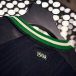 9xlAjqEDlZhJ6MW | UAE SPORT STORE Dark sports jersey on a hanger, featuring a green and white striped collar and the number 1908 printed on the back.