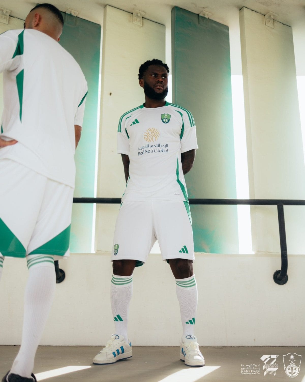 Two soccer players in white and green uniforms stand in a sunlit corridor, one facing forward with hands behind his back while a second player is partly visible on the left.