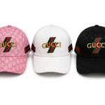 Three Gucci baseball caps in pink, white, and black, each featuring a gold Gucci logo and the signature red‑green stripe over a repeating GG pattern.