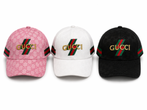 Three Gucci baseball caps in pink, white, and black, each featuring a gold Gucci logo and the signature red‑green stripe over a repeating GG pattern.