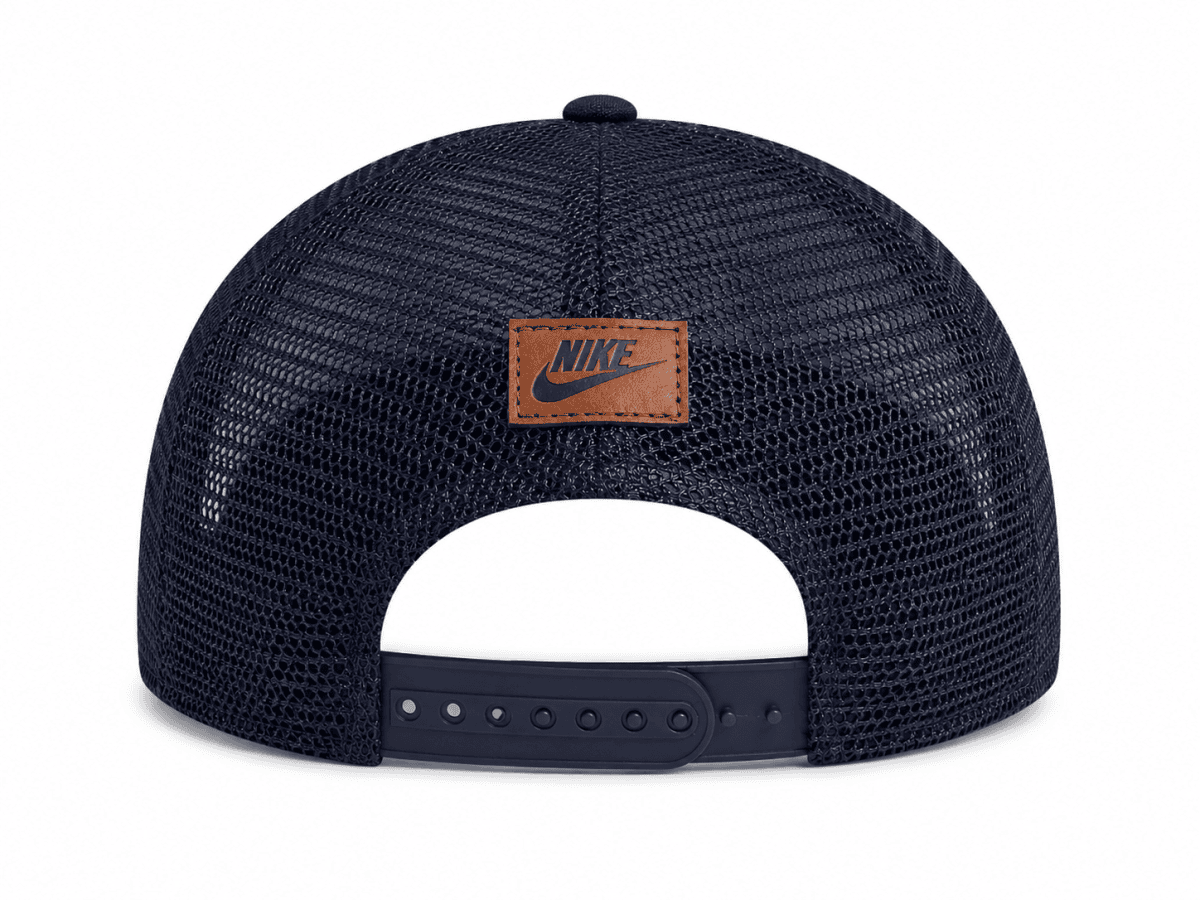 Rear view of a navy blue Nike trucker hat with mesh panels and a brown patch on the back. (Adjustable strap visible)