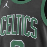 Dark gray Boston Celtics basketball jersey with green trim, large green-outlined 'CELTICS' letters across the chest and a white Jumpman logo near the shoulder, plus part of a green jersey number at the bottom.