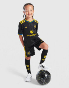 Young girl in a black soccer kit with yellow accents standing with one foot on a soccer ball, smiling at camera, brand patches visible (Snapdragon sponsor).
